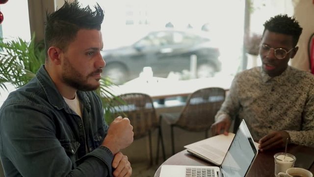 Serious multiethnic freelancers discussing work in cafe. Focused young men working with papers and laptop on coffee shop. Freelance concept