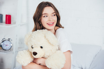 selective focus of smiling woman smiling and hugging teddy bear in bed
