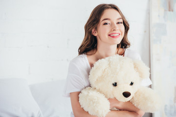 selective focus of happy woman smiling and hugging teddy bear in bed