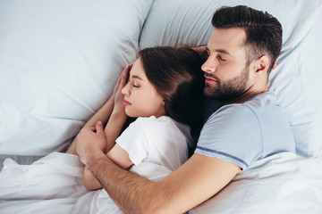 young couple sleeping on soft white bedding in bedroom while man embracing woman