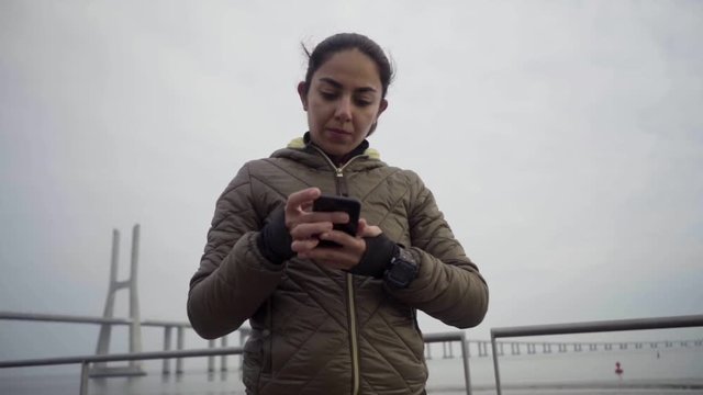 Serious sportswoman comparing time at smartphone and sport watch before running. Confident hindu woman with digital devices. Technology concept