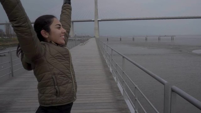 Smiling young woman running on wooden pier with raised hands. Successful female sportswoman outdoor during overcast day. Concept of winning 
