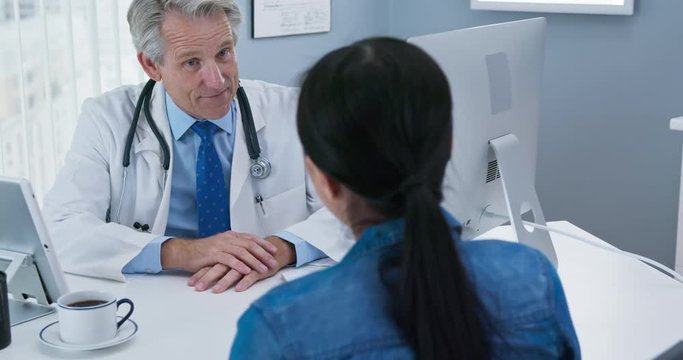 Older male doctor talking to female patient at his office desk