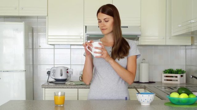 Young woman in pajamas enjoying early morning standing on kitchen drinking coffee. Sleepy female holding white cup with tea starting her day with energy trying to wake up
