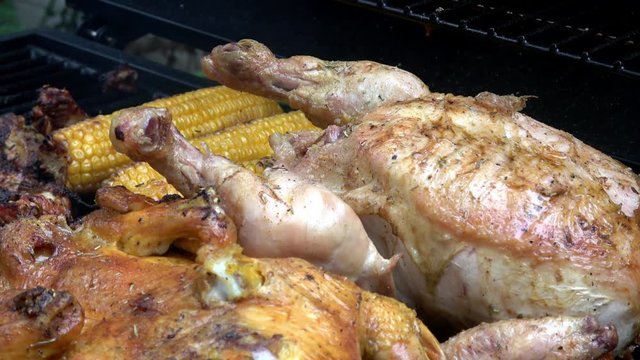 4K. Roast chicken on the barbecue grill. There are corn cobs and more meat around. Smoke comes out.