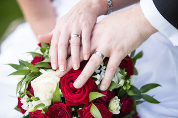 The bride and groom hold their hands with rings over a wedding bouquet with red and white flowers