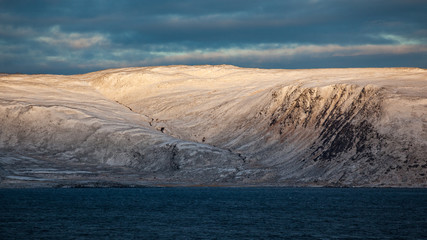 Snow covered rocks jut out from the sea. The cold evening sun goes down and the mountains shine in...