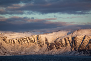 Snow covered rocks jut out from the sea. The cold evening sun goes down and the mountains shine in...