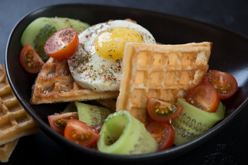 Close-up of waffles served with fried egg and fresh vegetables in a black bowl, selective focus
