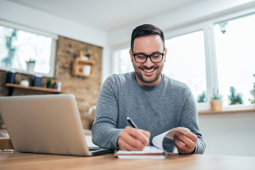 Low angle image of handsome man with laptop writing in notebook at home.