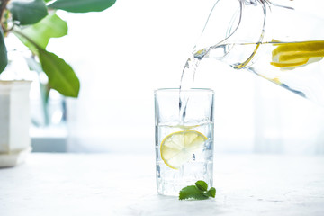 Pouring a glass of water with lemon, ice and mint on a white table © Natalya