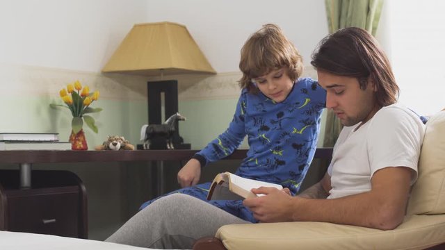 Two brothers sitting on arm chair, older brother read for small brother a book and they laughing to much