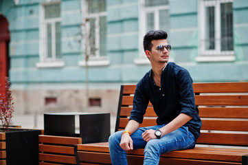 Stylish indian model man in casual clothes, black shirt and sunglasses posed outdoor at street of India and sitting on bench.