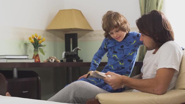 Older brother reading a book to his younger brother. Brothers in pajamas sitting in a chair discussing what they read in the morning