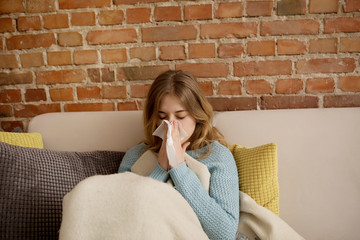Young sick woman siitng on sofa with running nose and tissues.
