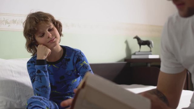 Older brother reading a younger brother's book sitting on the edge of the bed in the bedroom in the morning. Two brothers in pajamas having a good time together. Brothers relationship.