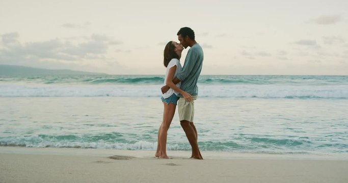 Happy young couple embracing and kissing on tropical beach at sunset, mixed race