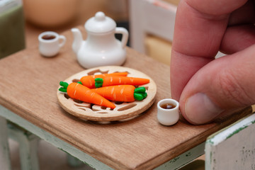 Small doll house table served for a tea party with tea pot, cups and plate full of carrots, A big hand holding a tiny cup with tea