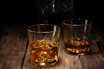 Two glass of whiskey with ice and crystal decanter on wooden table.