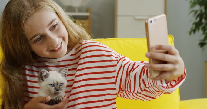 Portrait of the cute blond teen girl smiling and holding little kittycat while taking selfie photo on the smartphone camera in the nice room at home. Close up. Inside.