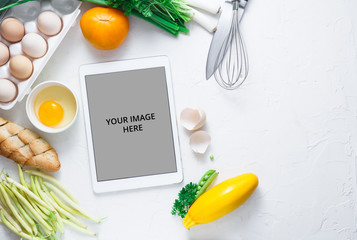 Digital touch screen tablet with fresh vegetables and kitchen utensils on background, top view