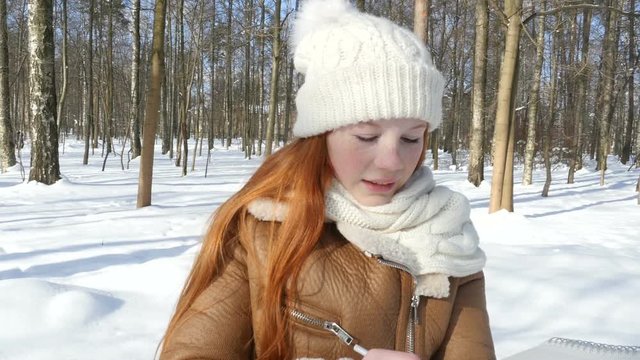 Red-haired teen child staying at winter park and draws in sketchbook. Beautiful little girl paints on paper