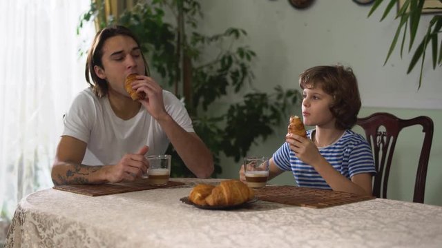 Two brothers sitting in kitchen table and eating croissant with cappuccino. Brothers relationship.