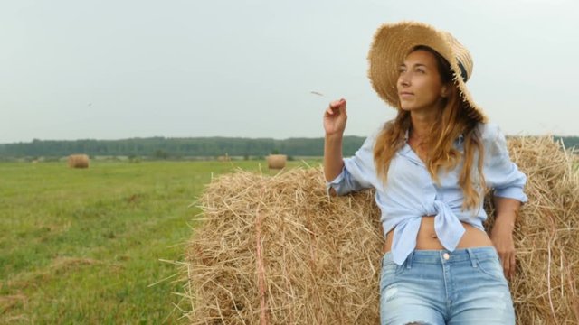 Pretty young woman stands near haystack and plays with straw. Throws the straws on camera. Farmer's wife walks on the field
