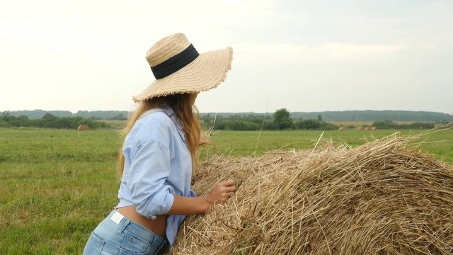 Pretty young woman stands near haystack and plays with straw. Farmer's wife walks on the field