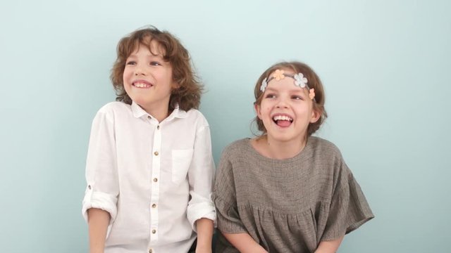 Children brother and sister on a light blue background in the studio laugh and look into the distance. Curly boy in a white shirt and a girl in a beige dress with a wreath on his head. Spring concept