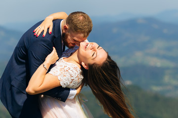 Newlyweds have fun at the top of the mountain. the bride has bend back and smiles and the wind blowing on her hair. the groom embraces the bride behind the waist