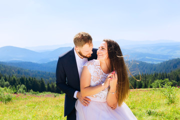 he groom embraces the bride behind the waist from the back and the bride closes her eyes and smiles genuinely. beautiful mountains covered with forests