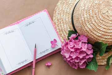 Obraz na płótnie Canvas Female open notebook with pink pen, hydrangea flowers and straw hat. Romantic workplace for dreaming, writing goals, woman freelance. Flat lay in vintage style.
