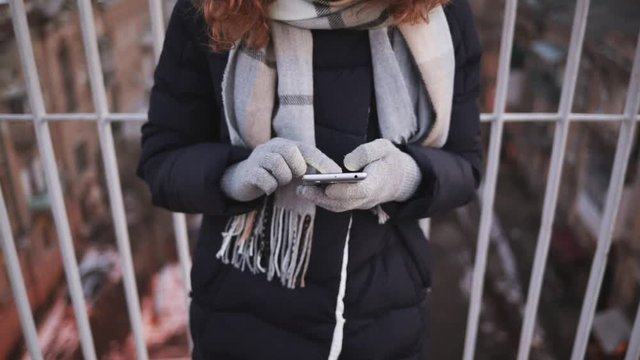 Young woman wearing gloves uses smart phone outdoors in city in winter, close-up.