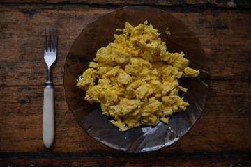 Scrambled eggs in a plate next to a fork on the background of wood
