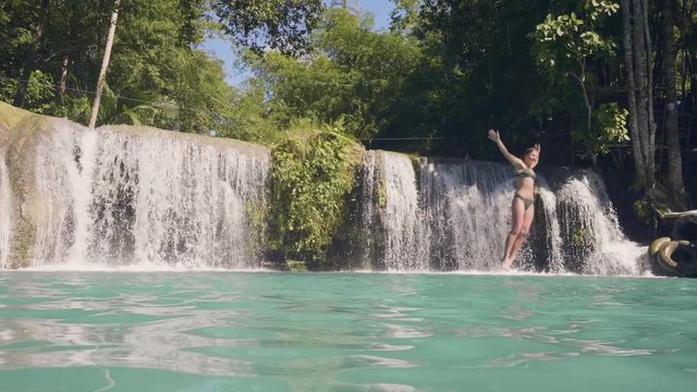 Young woman jumping in waterfall lake from rope and stick slow motion. Cheerful woman jumping from rope in blue water from flowing waterfall in rainforest.