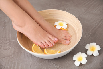 Obraz na płótnie Canvas Woman soaking her feet in bowl with water, orange slices and flower on grey background, closeup. Spa treatment