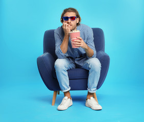 Fototapeta premium Emotional man with 3D glasses and popcorn sitting in armchair during cinema show on color background