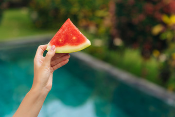 Close up slice of watermelon in female hand on background of blue swimming pool and greenery