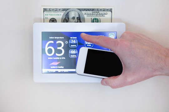 Male hand holding smart phone to operate heating or cooling via digital touch screen thermostat for home energy savings