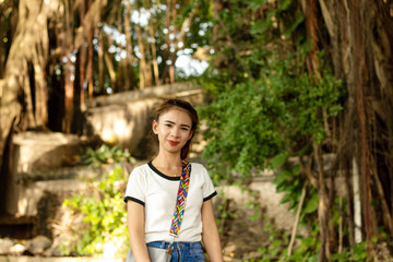 Asian woman with brown hair A small woman White skin, red lips Wearing a white shirt Wearing blue jeans Standing smile Gray shoulder bag With rocks and large trees on the back in the park