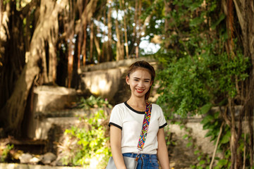 Naklejka na ściany i meble Asian woman with brown hair A small woman White skin, red lips Wearing a white shirt Wearing blue jeans Standing smile Gray shoulder bag With rocks and large trees on the back in the park