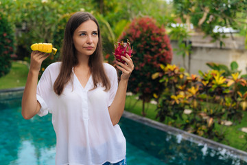 Portrait of cute girl with pensive face holds mango and dragon fruit, pitaya in her hands