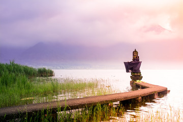 Small Hindu Temple on The Lake Side With Connecting Bridge During Sunrise in Bali on Cloudy and Misty Day with Mountain as Background