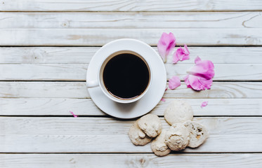 Cup of coffee and beautiful petals of peonies and biscuit on white wooden background. Copy space.