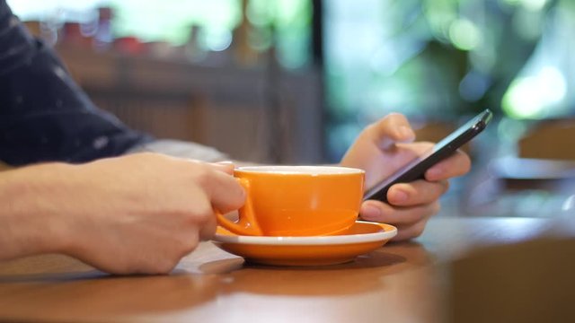 Close-up of hand of a man holding and using smart phone while drinking from a cup of steaming coffee in a modern coffee shop in Medellin, Colombia, Latin America. High quality 4K ProRes 422 HQ