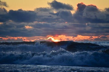 Storm on the sea at sunset. Blue sky with dark clouds. The light of the sun on the horizon. Big waves roll ashore. Splashes and mist over the wave.