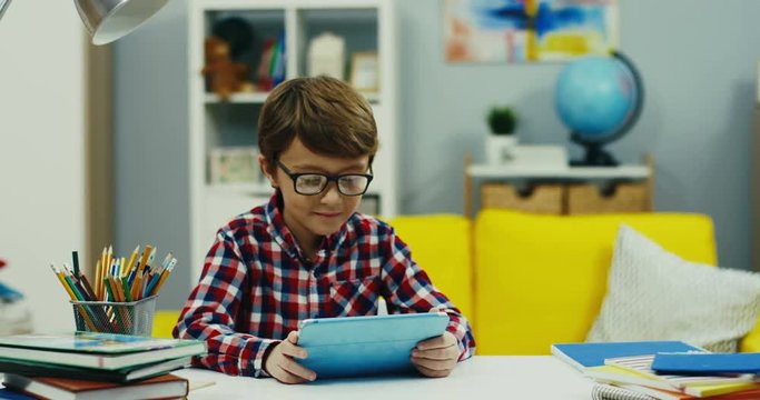 Camera zooming in of the small teen boy in glasses and plaid shirt watching something on the tablet device at the desk in the cozy kid's room.