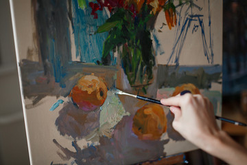 A young woman artist sits in a bright studio on a blue chair and paints a picture in oil on the easel.