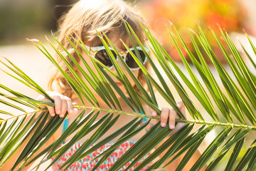 Closeup portrait of adorable little girl at the tropical resort, hiding behind the palm at the sunny summer day